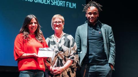 Three people on stage, in front of a screen. The woman on the right is wearing a red top, jeans, is holding a clapper board prize and has long dark hair. The woman in the middle has short hair, and a black and white dress on and the man to the left is wearing a T-shirt, jacket and trousers. 