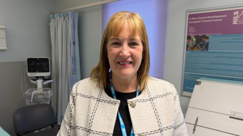 A woman wearing a light-coloured, checked jacket and an NHS lanyard stands in a hospital room. Medical equipment, a curtained area and information boards are visible in the background.