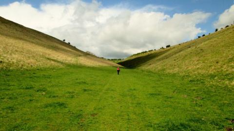 A solitary hiker walks through a grass-lined valley on the Yorkshire Wolds Way near Holm Dale