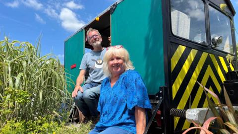 Adrian and his wife sat on the footplate and entrance to his train surrounded by plants. Both are smiling at the camera