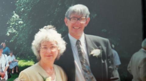 An old colour photo of Jean Turnbull and Raymond Turnbull, taken outside at a wedding or similar event. They both have large glasses and flowers in their button holes. Jean has white-grey hair to just above the shoulder and is wearing a yellowish tan light jacket. Raymond has grey hair brushed forward and wears a dark grey suit jacket over a very pale blue shirt and patterned tie. It is a sunny day and a large tree thick with leaves forms the background. A few other guests can be seen milling around.