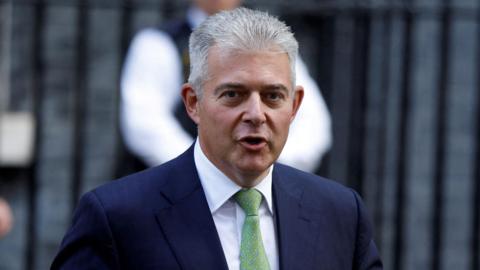 Brandon Lewis walking down Dowing Street carrying a folder under his arm. He is looking at the camera and saying something. He is wearing a suit and tie. Behind him out of focus is a man in police uniform and and another man wearing a suit and carrying a folder. Behind them the black railings and bricks of Downing Street buildings.