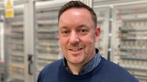 A man smiles at the camera. He has short brown hair and stubble. He is wearing a dark blue collared shirt and dark blue jumper. He is standing in front of glass fronted metal cabinets which have batteries on charge inside.