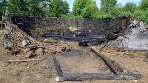 Wattle and daub walls of an Iron Age replica roundhouse with a pile of sticks and burnt wooden frame of an entrance and piles of ash and thatch.