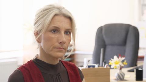 A woman with blonde hair sits in front of a desk. There is a large black chair in the background and a small vase with flowers on the desk.