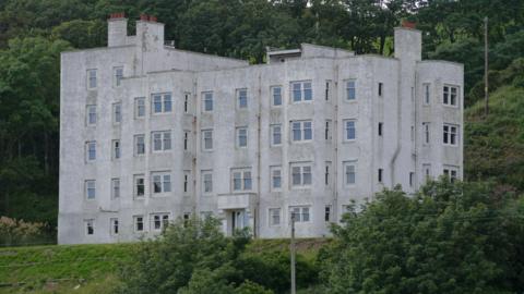 A large five storey white building, once a hotel and now in a ruined state. It sits among trees and greenery.