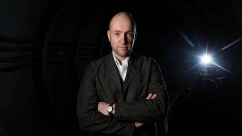 Derren Brown. He is pictured with his arms folded, looking straight at the camera with a slight smile. He is bald and has some stubble on his chin. He is wearing a dark grey jacket, a white shirt and a watch.