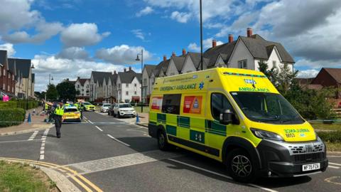 An ambulance parked in front of a police cordon as an officer directs traffic away
