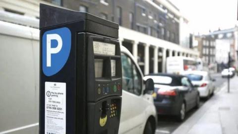 The side of a black parking metre. There is a blue sticker with a white P on it. In the background is a white van parked behind a number of cars in a street