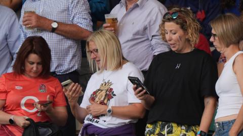 Women watching the New Zealand vs Ireland Women's rugby world cup match look at their phones during the test on an emergency alert system.