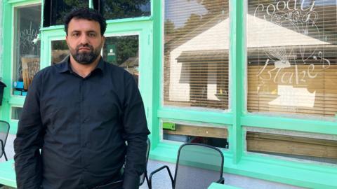 Shwan Khder is wearing a black shirt. He has a beard and looks unhappy. Behind him there is a building with large windows and light green window frames.
© BBC