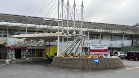 The accident and emergency department at the hospital. It is a concrete building with a canopy out front. A concrete roundabout is planted with many colourful flowers. 