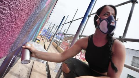 A woman wearing a black vest and black ripped jeans and a face mask. She has long black hair tied into a plait and is kneeling on scaffolding, using a spray can to paint the side of a building.