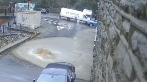 Two cars and a van close to a large pool of brown water covering a roadway next to a steep stone wall