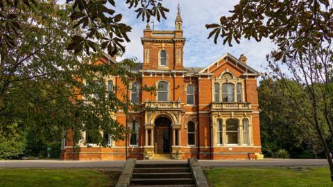 An exterior view of Weelsby Hall in Grimsby. The Victorian mansion house has been constructed using red bricks and yellow coloured sandstone. A lawn and stone steps are in the foreground and the house is surrounded by grassed areas and large trees.