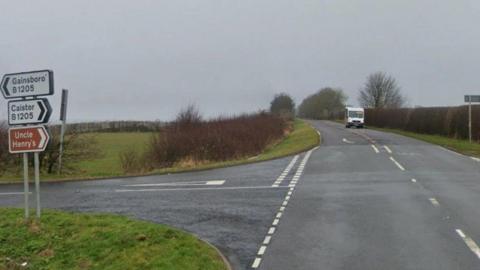 Google street view image of a country road junction. In the distance is a white van driving towards the camera. On the left are three road signs stacked in a column. The first sign reads 'Gainsborough B1205' with an arrow pointing to the left, the second reads 'Caistor B1205' with arrow pointing to right, a third is a brown sign reading 'Uncle Hentry's' with an arrow pointing to the right.