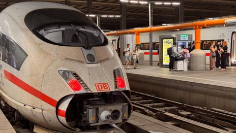A DB Deutsche Bahn passenger train is seen at a standstill as passengers with suitcases wait on the platform to the right.