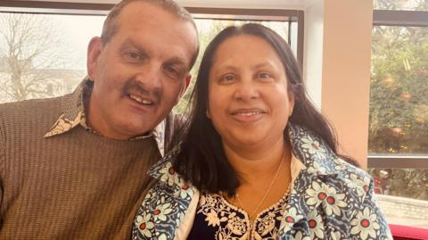 A man with a moustache and a brown sweater with a shirt collar over the top of it and a woman with dark hair and wearing a flower-patterned coat and a flower-patterned top.