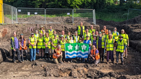 A group of volunteers in yellow hi-vis jackets standing in the dig site. They are wearing hi vis jackets and are holding a Cumberland flag.