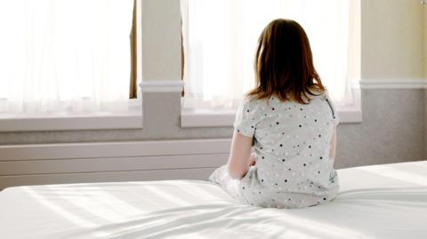 A girl wearing polka dot pyjamas, sat on the edge of a bed with a white duvet. She is looking out towards two windows with white curtains. 