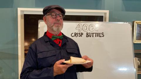 A man in a blue lab coat holds a crisp butty next to a sign reading "400 crisp butties".