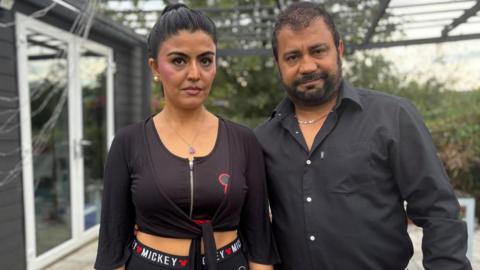 A woman and a man stand on a garden patio looking straight at the camera. They are both wearing black.
