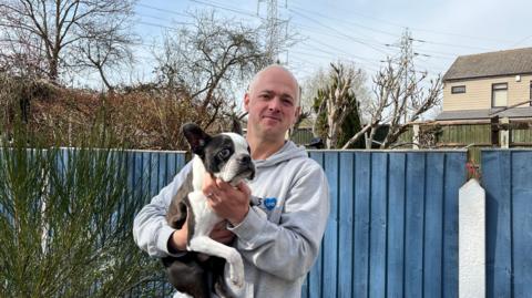 A man in a grey hoodie holds a small black and white dog in his garden, with two pylons in the background behind him.