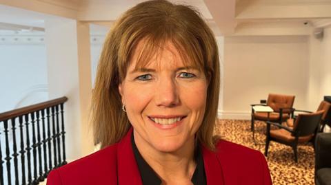 A woman with shoulder-length blonde hair is smiling at the camera. She is standing on what appears to be an indoor balcony. On the left is a railing looking onto a bigger room. On the right are chairs and a table for people to sit and chat. There is yellow and brown carpet on the floor and the walls and ceiling are white. She is earing a dark top under a red jacket.