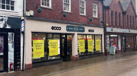 A cream coloured shop front with a red brick exterior above is pictured on a high street on a wet day. The store has a green sign that reads Robin Elt Shoes. 