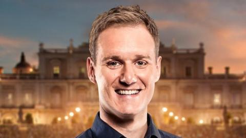 A man stands in front of a palace. He has short blonde hair and is smiling at the camera.