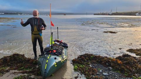 David Higson, who is a middle-aged man, wearing a wetsuit and smiling with his thumbs up. He is standing next to his raft at Port St Mary Bay.