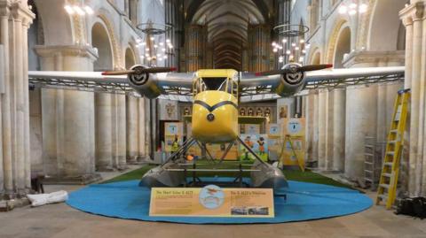 A yellow floatplane with silver wings and propellers. It is on a blue, circular carpet inside the middle of a cathedral.