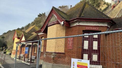 A boarded up brown brick building at the base of a cliff