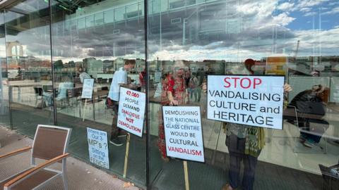 Campaigners seen through the glass front of the building with multiple placards. The first on the right reads 'Stop vandalising culture and heritage', the next 'Demolishing the National Glass Centre would be cultural vandalism', then to the left there is '47,000 people demand dialogue not demolition' and finally on the far left 'Where are our 3 MP's? Sitting on the fence.' Due to the reflection it is hard to see how many people are inside, but it looks to be at least 10. Cranes, buildings and clouds are reflected in the glass.