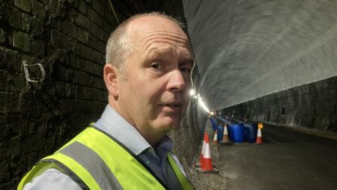 Man with very short hair wearing hi-viz jacket stands with tunnel in the background