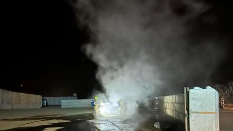 Smoke bellowing from a vehicle fire outdoors with two firefighters stood either side with hoses. There is a large area with walls on the left and right. The vehicle is central and the ground around it is wet. 