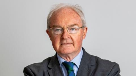 A man with white hair looking at the camera, he has glasses and dark blazer, blue shirt and green and blue tie