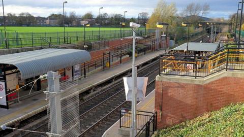 A picture of the Metro station. It is outdoors and has a curved metal roof on one platform. The opposite platform has a flat roof and there are stairs leading down to it.