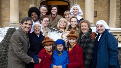 Cast members of Call the Midwife dressed in period clothing stand on a set of snowy steps holding a clapper board.