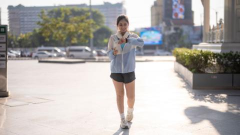 Woman wearing a rain coat and gym shorts walking whilst checking the time on her watch.