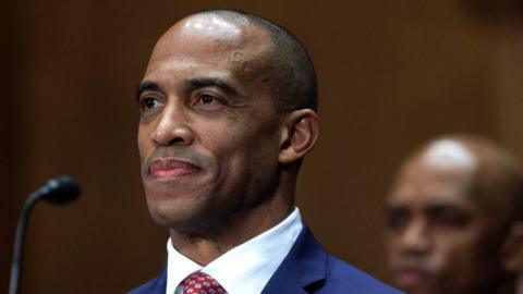 Scott Turner seen in the US Capitol. He is sitting at a microphone and wearing a blue suit with a red spotted tie. He has a slight smile on his face. 