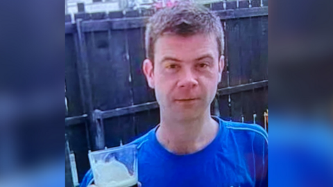 Mark Dorrian is wearing a bright blue t-shirt and has short brown hair. He is looking directly at the camera. 