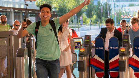 A man wearing a green shirt and dark jeans. He is holding a selfie stick in his right hand and his left hand is in the air. He appears to be running with a big grin on his face. Behind him are barriers and several people standing behind them