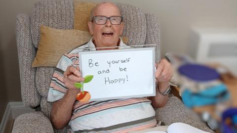 A bald man wearing black glasses and a stripy polo shirt. He's sitting on a light brown armchair and holding up a whiteboard with "be yourself and be happy" written on it. He's smiling at the camera.