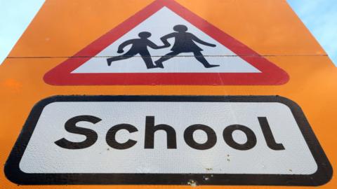 A picture of a yellow sign. It says the word school on it with a graphic above it.