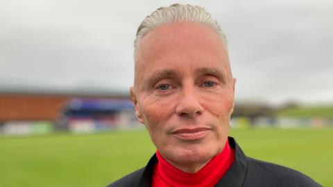 Barrie Drewitt-Barlow has blond, slicked back hair. He is wearing a black suit jacket over a red roll-neck top. He looks serious while standing in front of the pitch at Maldon and Tiptree Football Club.