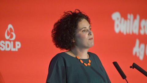 Claire Hanna. She has dark curly hair, a dark green top and orange necklace. She is standing in front of microphones. A red background is behind her with SDLP branding on it.