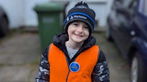 Harlee is smiling at the camera. He is wearing a black and dark blue hat, with a grey and black camo coat, and an orange hi-vis jacket.