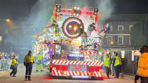 A carnival cart makes its way down a road, flanked by people in hi-vis vests.