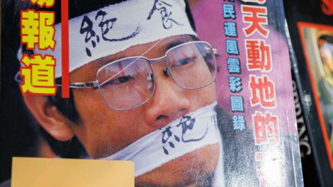 Close-up of publication cover showing Chinese man wearing wire-rimmed glases, a headband reading "hunger strike" in Chinese and matching white cloth gag tied across his mouth - the book is a photo chronicle of the Tiananmen Square democracy movement